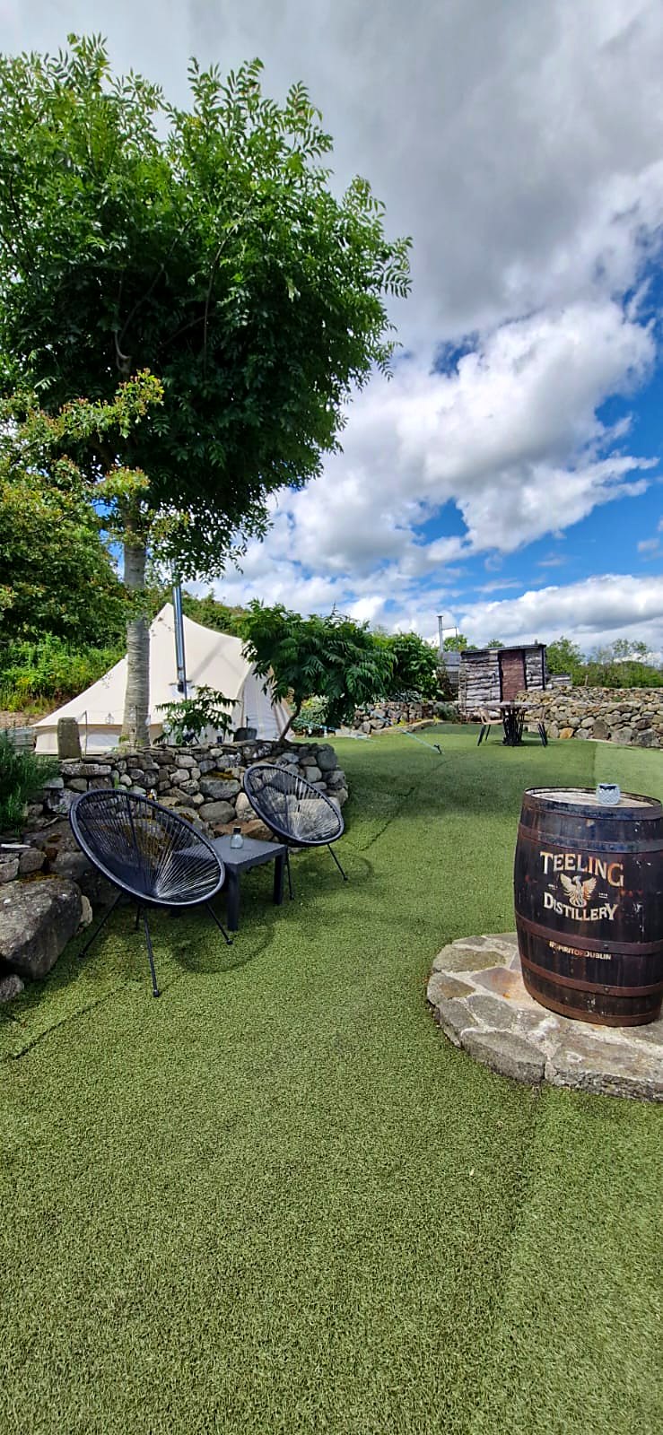 Two chairs and a low table under a tree on the lawn at Fairy Fort, with an old barrel on a stone circle