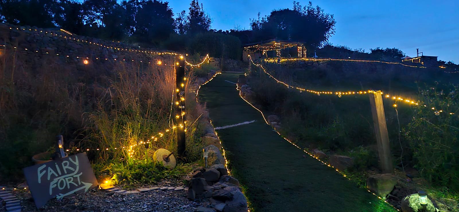 The lit path up to Fairy Fort at dusk, a chalk sign reading Fairy Fort beside stone walls strung with lights