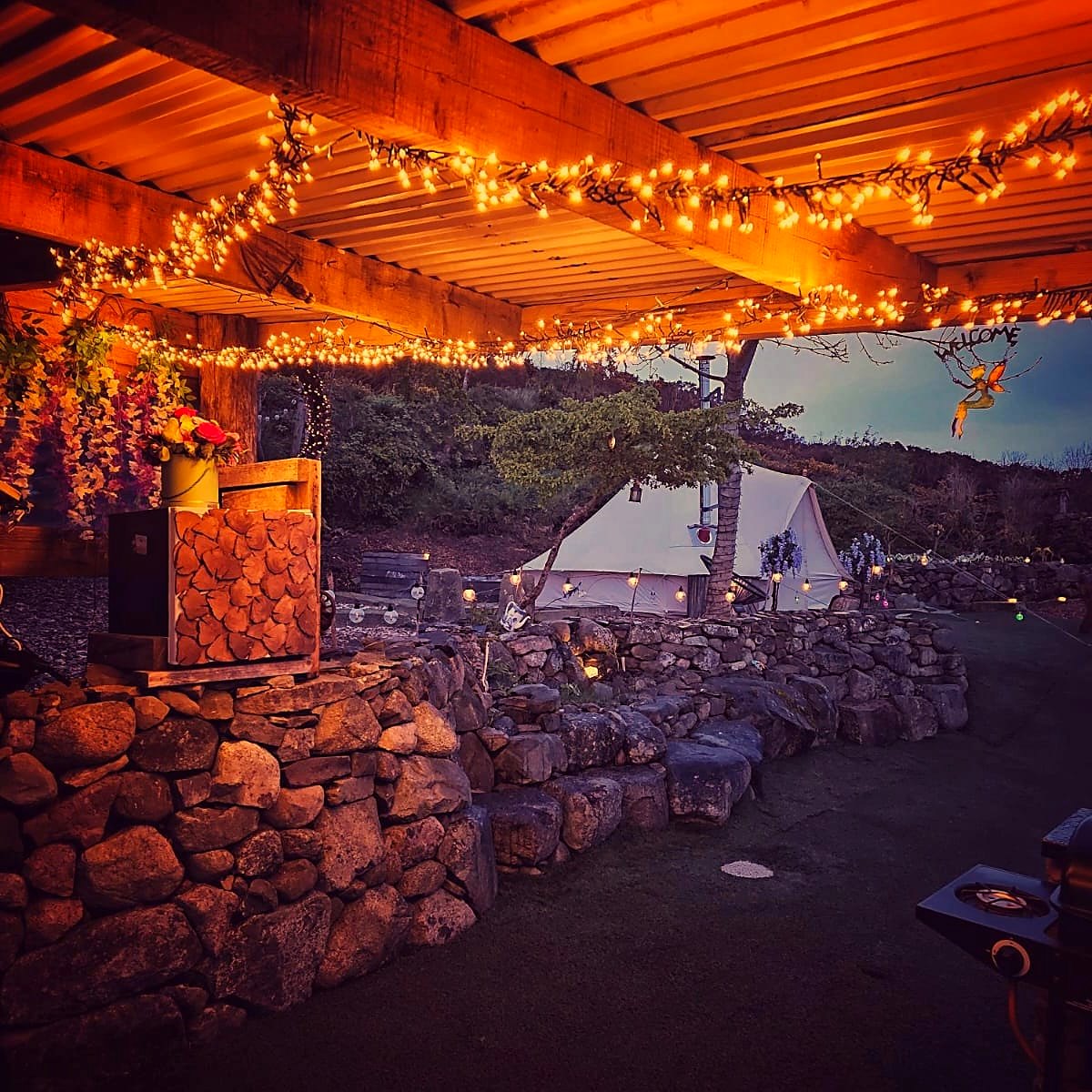 Covered shelter at Fairy Fort strung with fairy lights at dusk, the lit bell tent beyond the stone wall