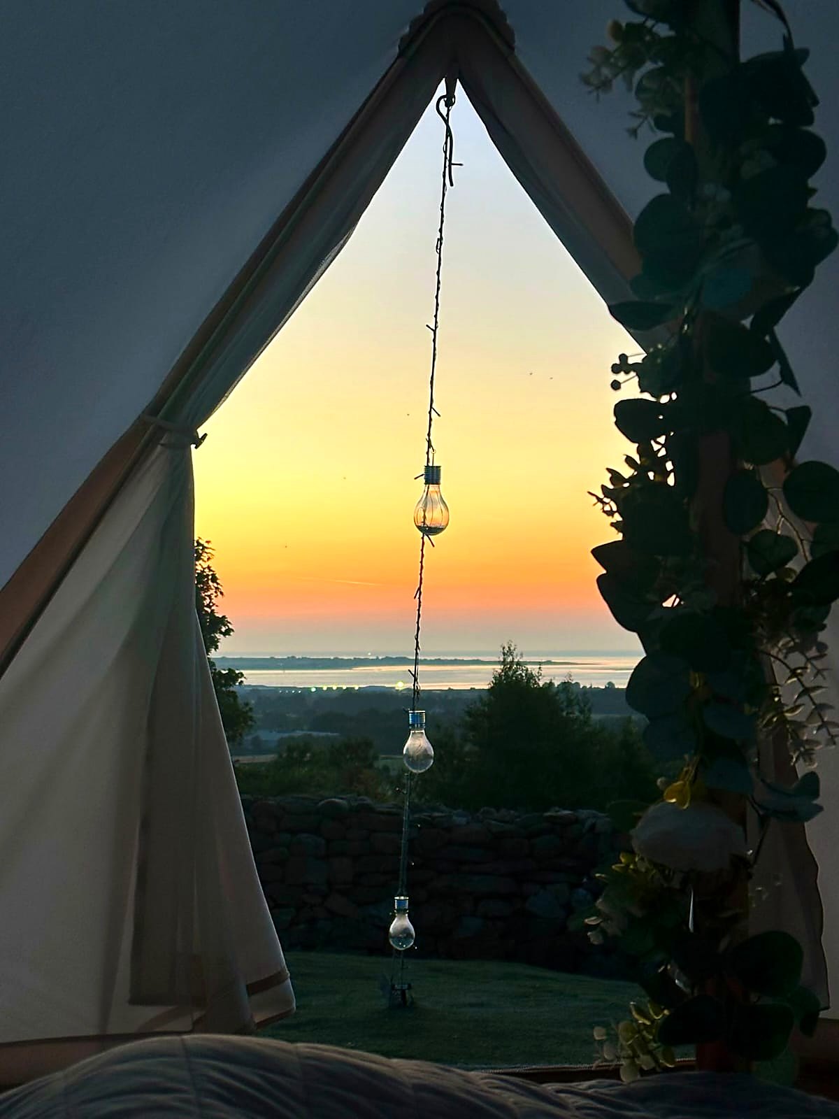 Sunset over Carlingford Lough framed by the open doorway of Fairy Hill bell tent, a hanging bulb and greenery at the edge